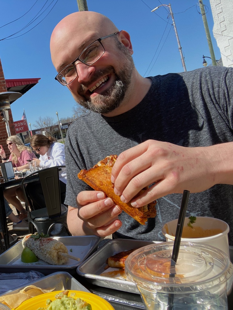 Author outside at a table with a birria taco in his hand. Elote also present on the table in front of him. He has a beard and glasses and is smiling.